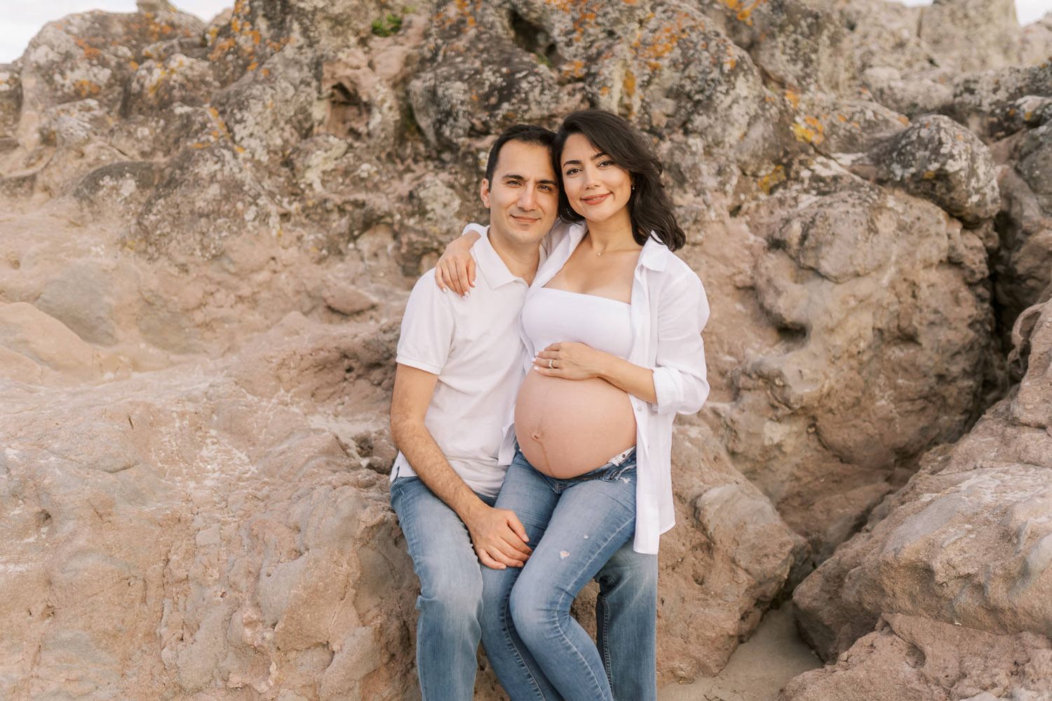Happy couple posing outdoors near rocky cliffs, woman with pregnant belly embracing man in white shirt and jeans.