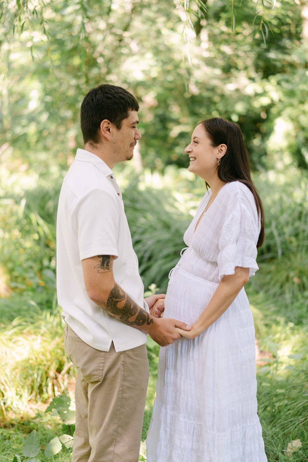 A couple shares a tender moment outdoors, the woman in a white dress cradling her pregnant belly while her tattooed partner holds her hands.
