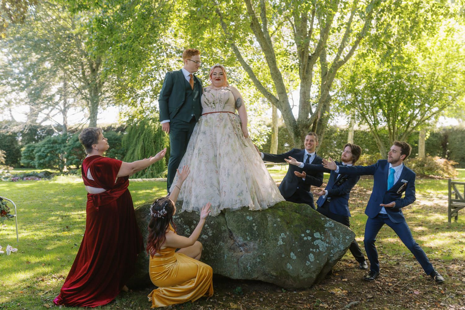 Wedding couple stands on a large rock surrounded by colorful bridesmaids and groomsmen in a lush garden setting.