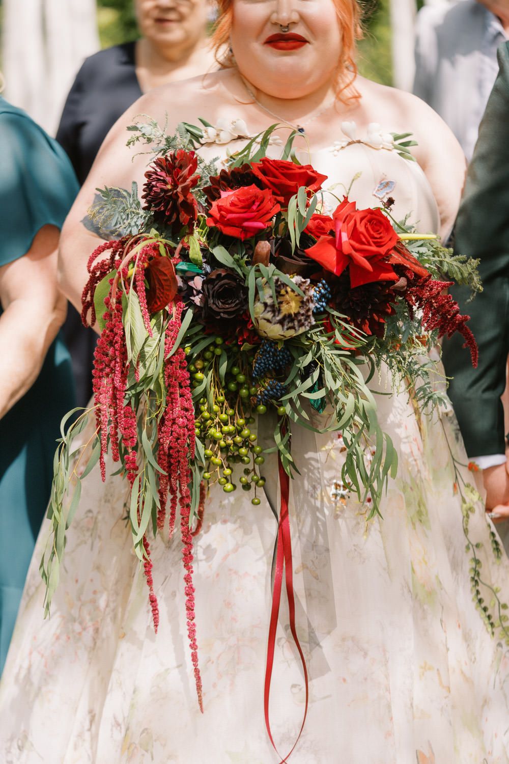 Bride holding a lush cascading wedding bouquet with red roses, amaranthus, and greenery, wearing a floral gown.