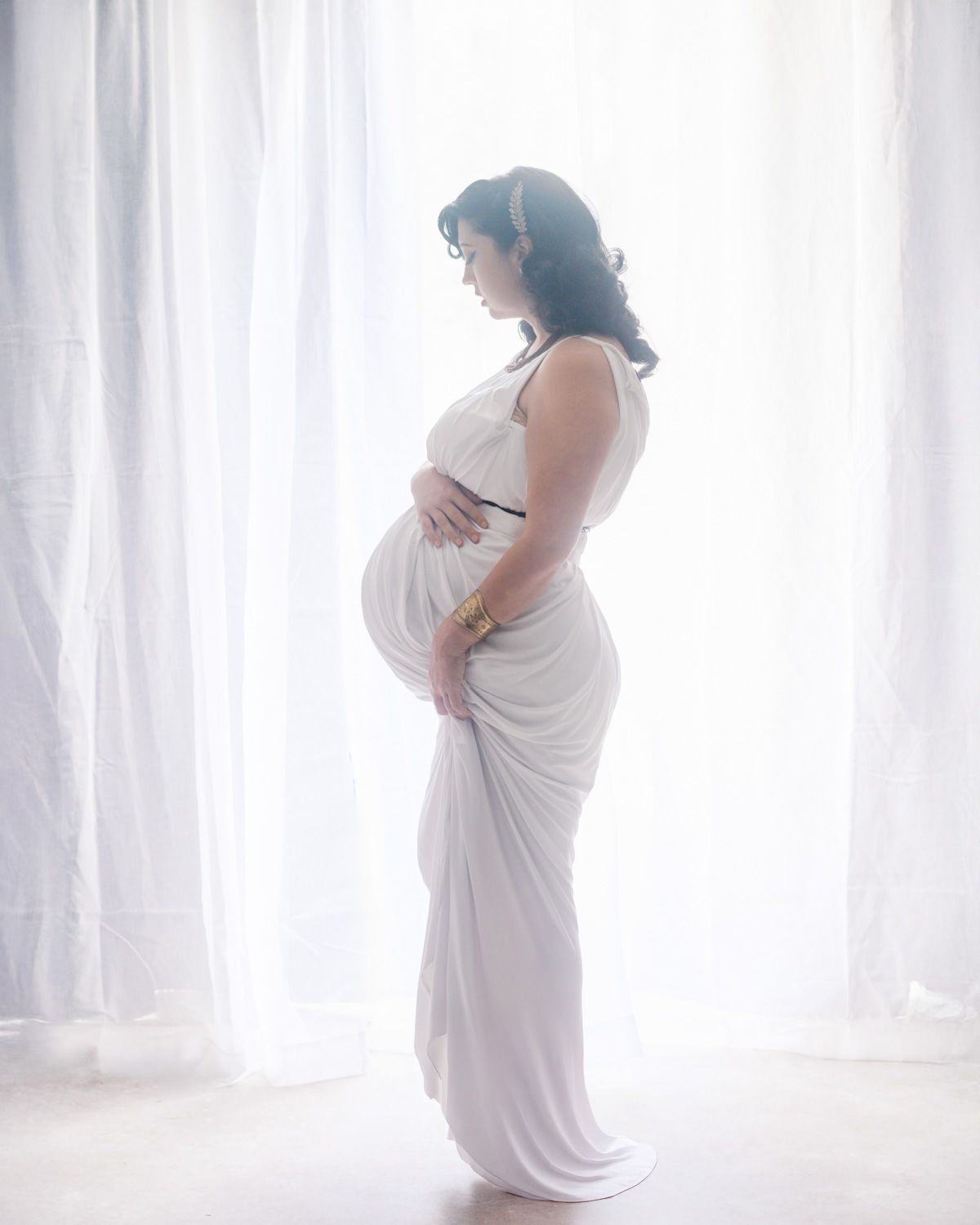 Pregnant woman in white draped gown stands sideways by sheer curtains, cradling her baby bump in a serene maternity portrait.