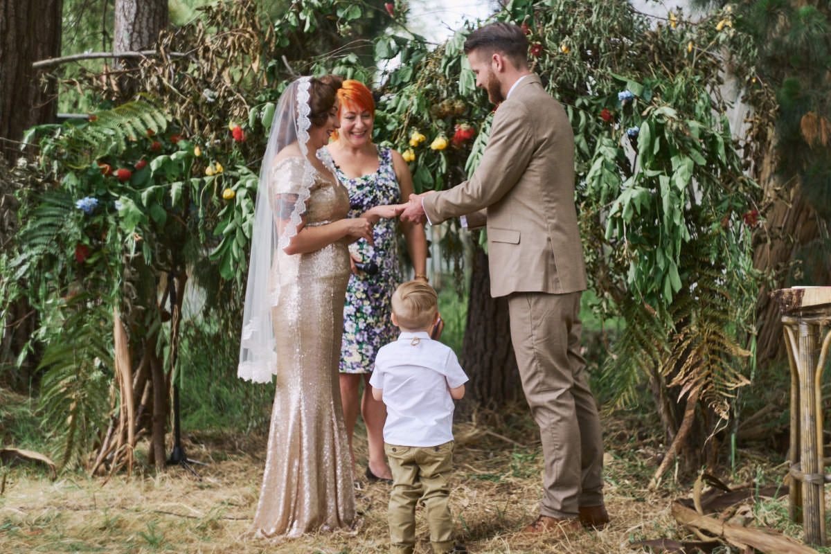 Couple exchanging vows outdoors under lush green arch with officiant and child present at rustic woodland wedding ceremony.