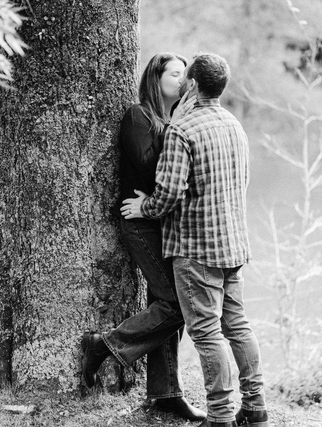 Black and white photo of a couple sharing a romantic kiss against a large tree in a natural outdoor setting.