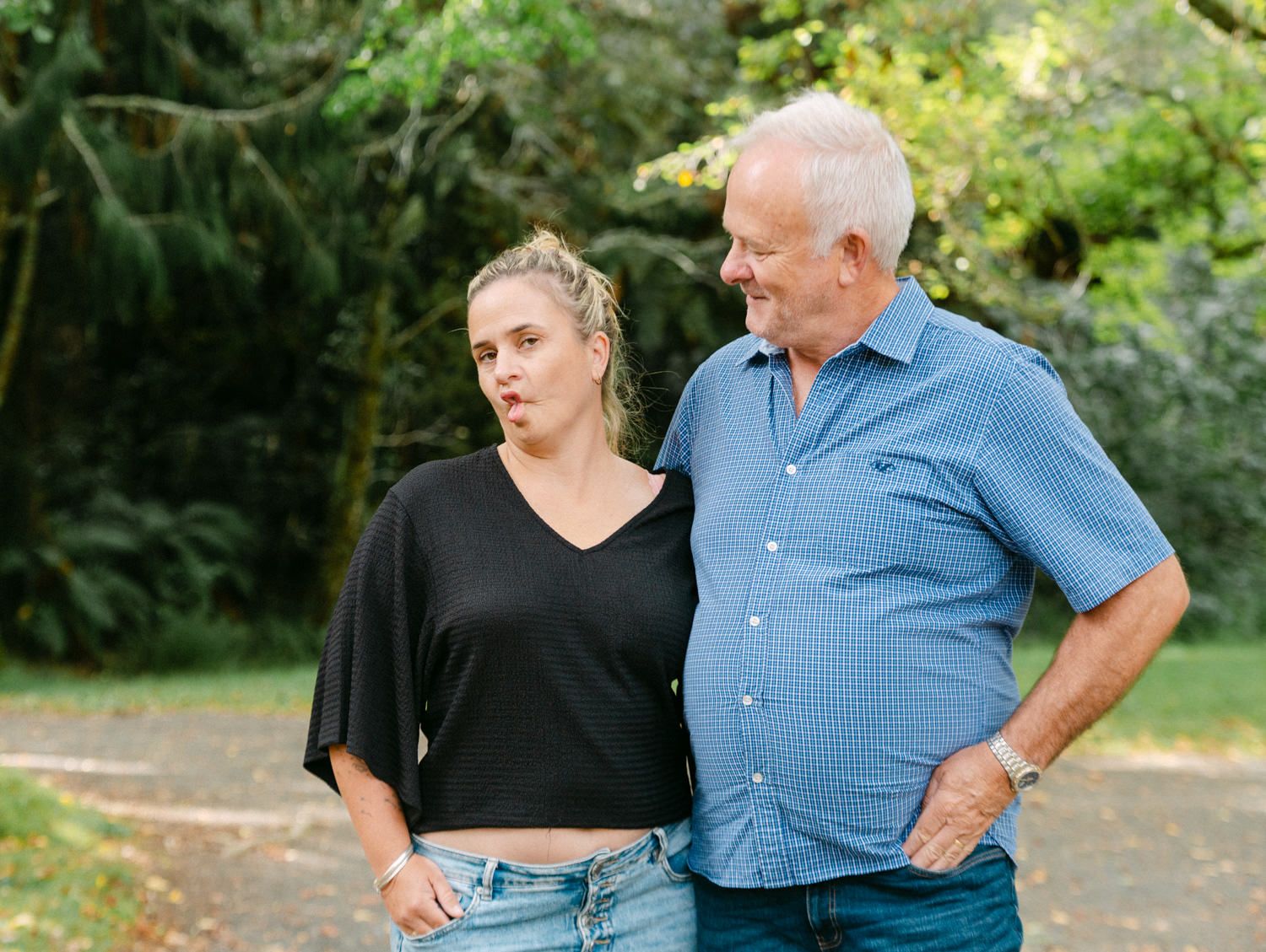 A young woman and an older man stand together outdoors, smiling, surrounded by lush green trees.