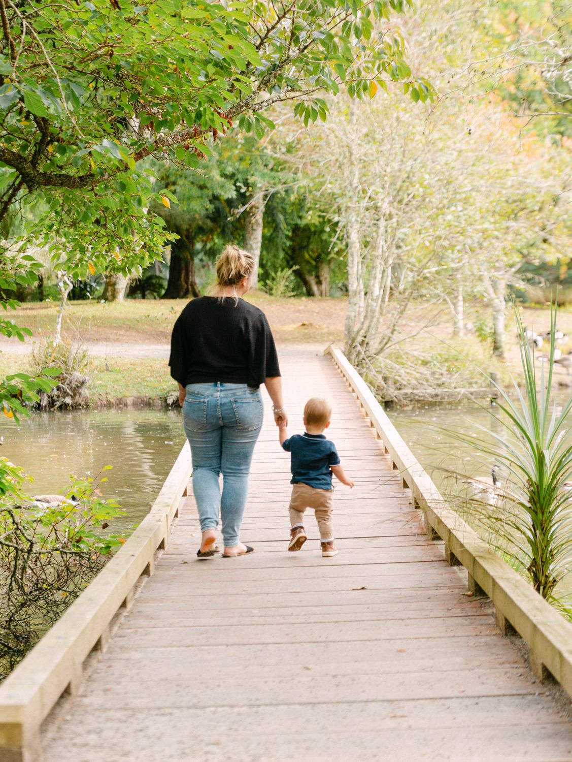 A mother and toddler walk hand-in-hand across a wooden bridge surrounded by lush green trees and a calm pond.