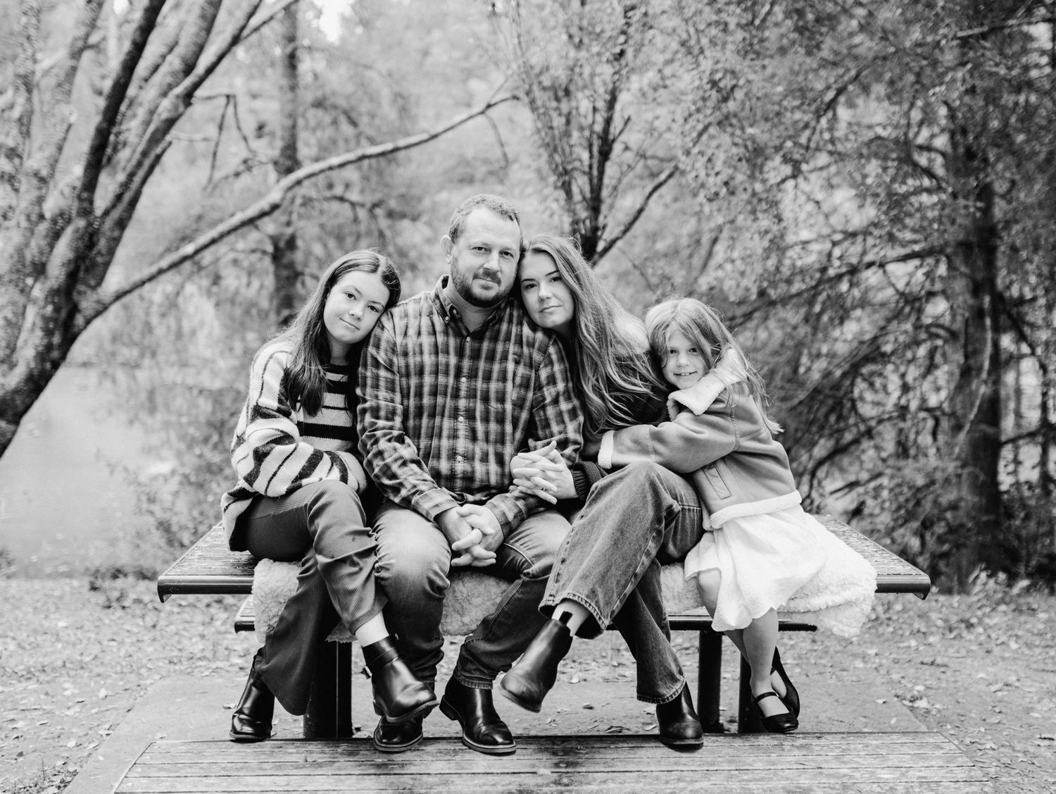 Black and white family portrait of parents and two daughters sitting on a bench surrounded by autumn trees.