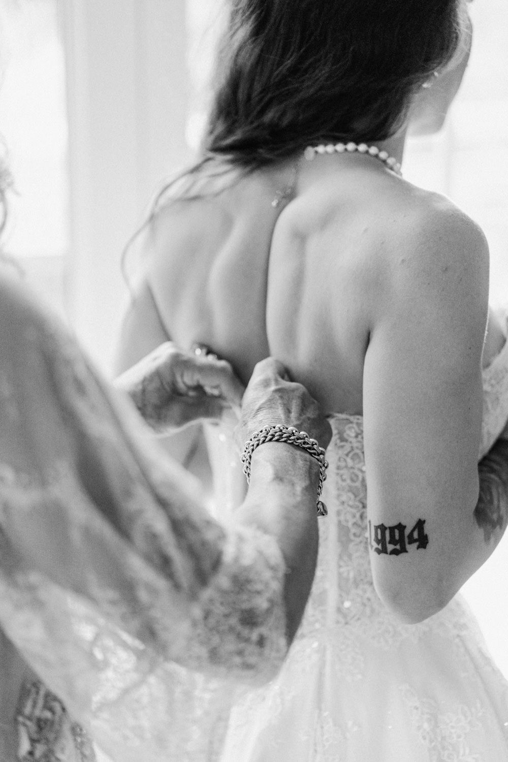 Black and white photo of bride in lace wedding dress being buttoned up, assistant with chain bracelet and 1994 tattoo visible.