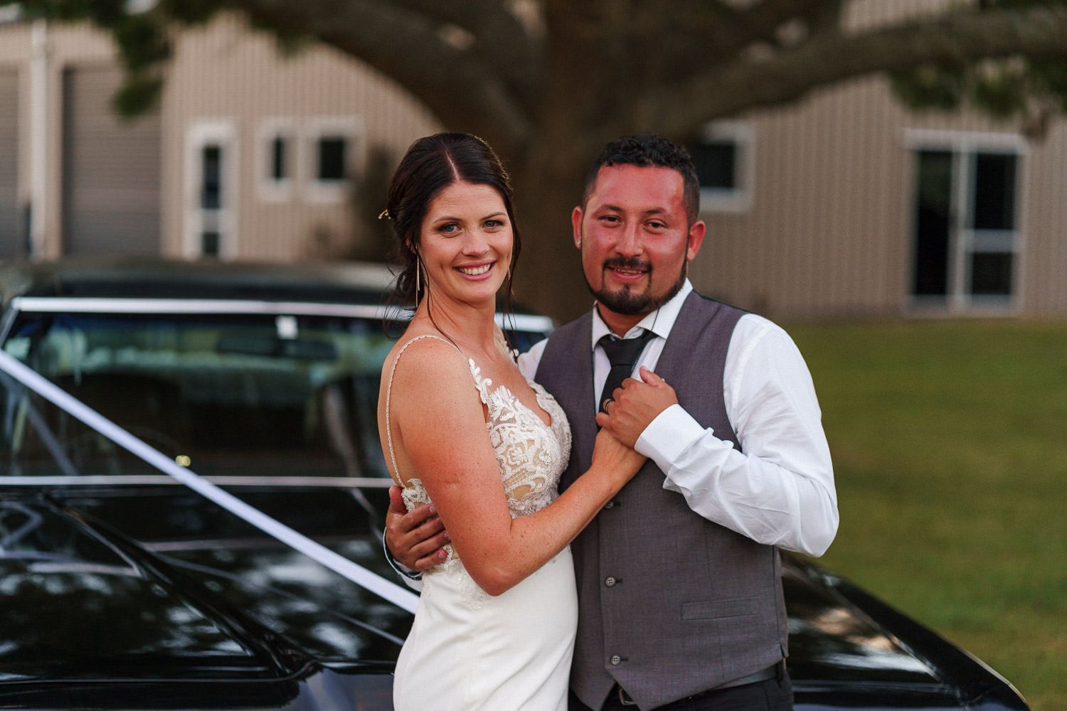 Bride and groom posing elegantly in front of a classic black car at their outdoor wedding venue.