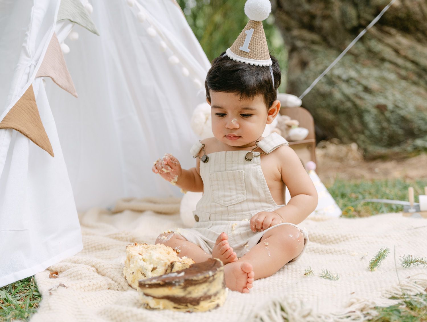 Baby in linen romper wearing birthday hat smashes first cake outdoors near white teepee on blanket.