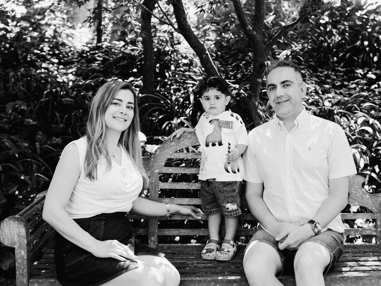 Black and white family portrait of parents and toddler son sitting on a wooden bench surrounded by lush greenery.