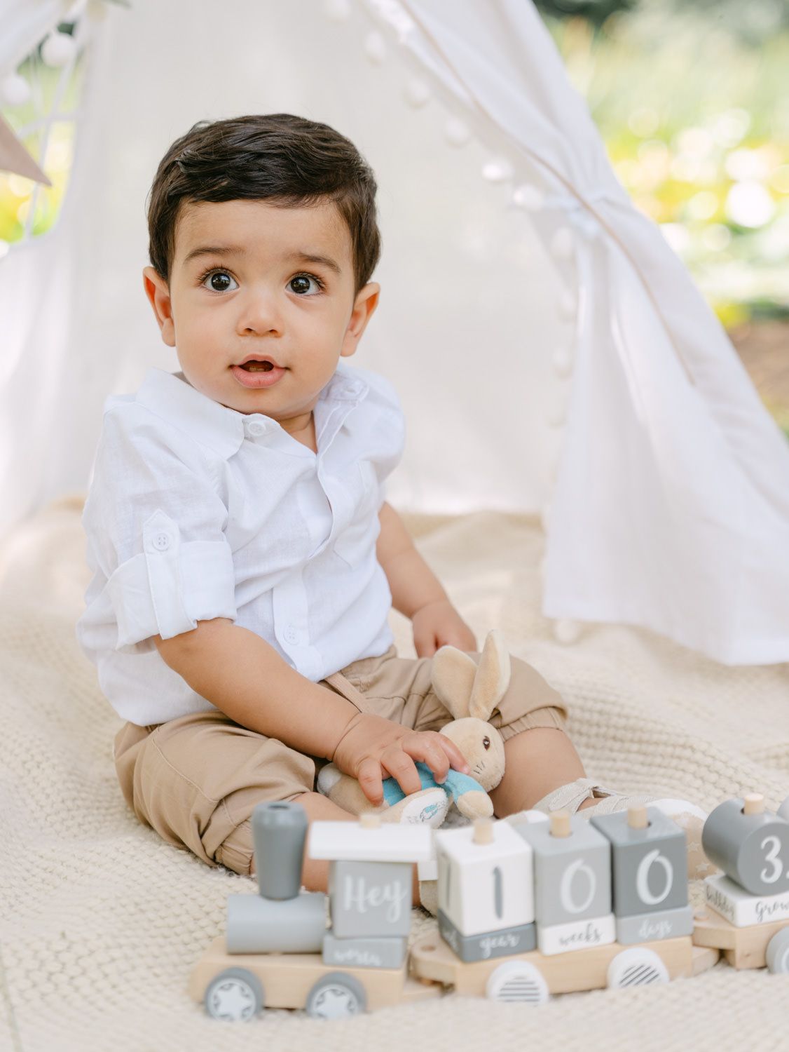 A baby boy in a white shirt sits on a blanket near a teepee, playing with wooden toy blocks and a train.