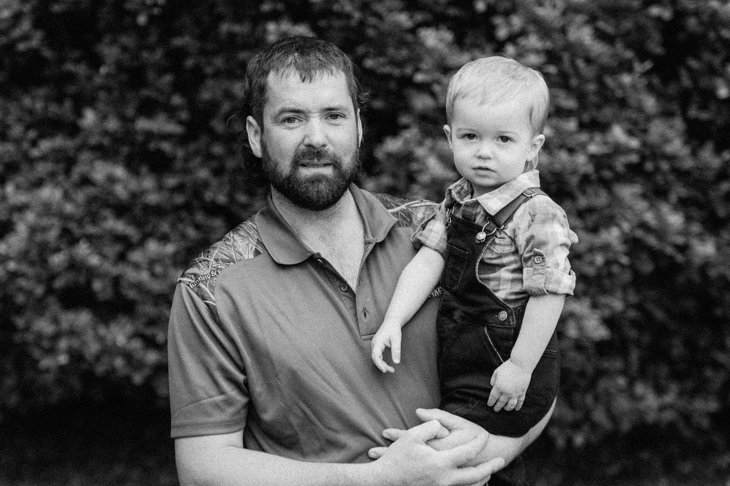 Black and white portrait of a bearded man holding a toddler boy outdoors with lush green foliage background.
