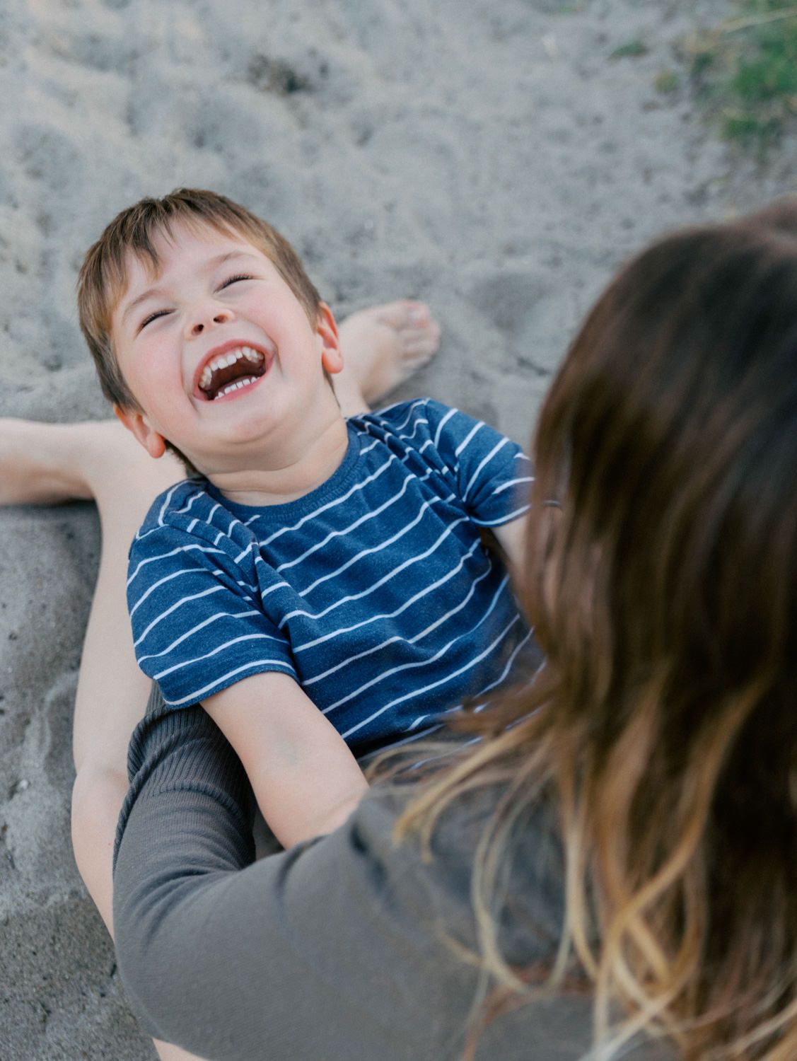 A laughing young boy in a blue striped shirt plays in the sand with a woman during a joyful beach outing.