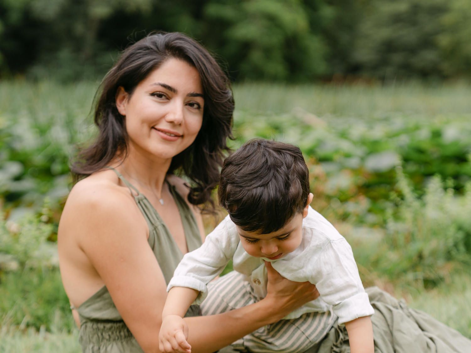 A woman in a green dress sits outdoors holding a young boy in a lush green garden setting.