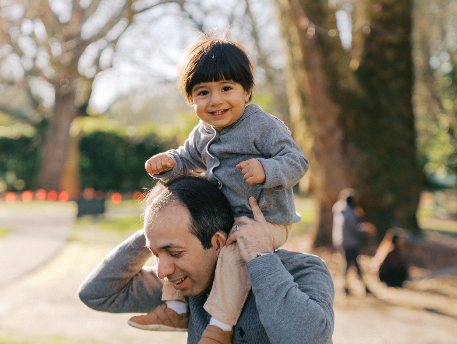 A smiling toddler sits on a man's shoulders in a sunny park, both enjoying a playful outdoor moment together.