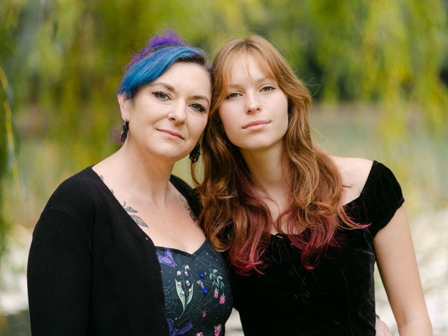 Two women with colorful hair pose together outdoors near green trees, one with blue hair and one with auburn and pink highlights.