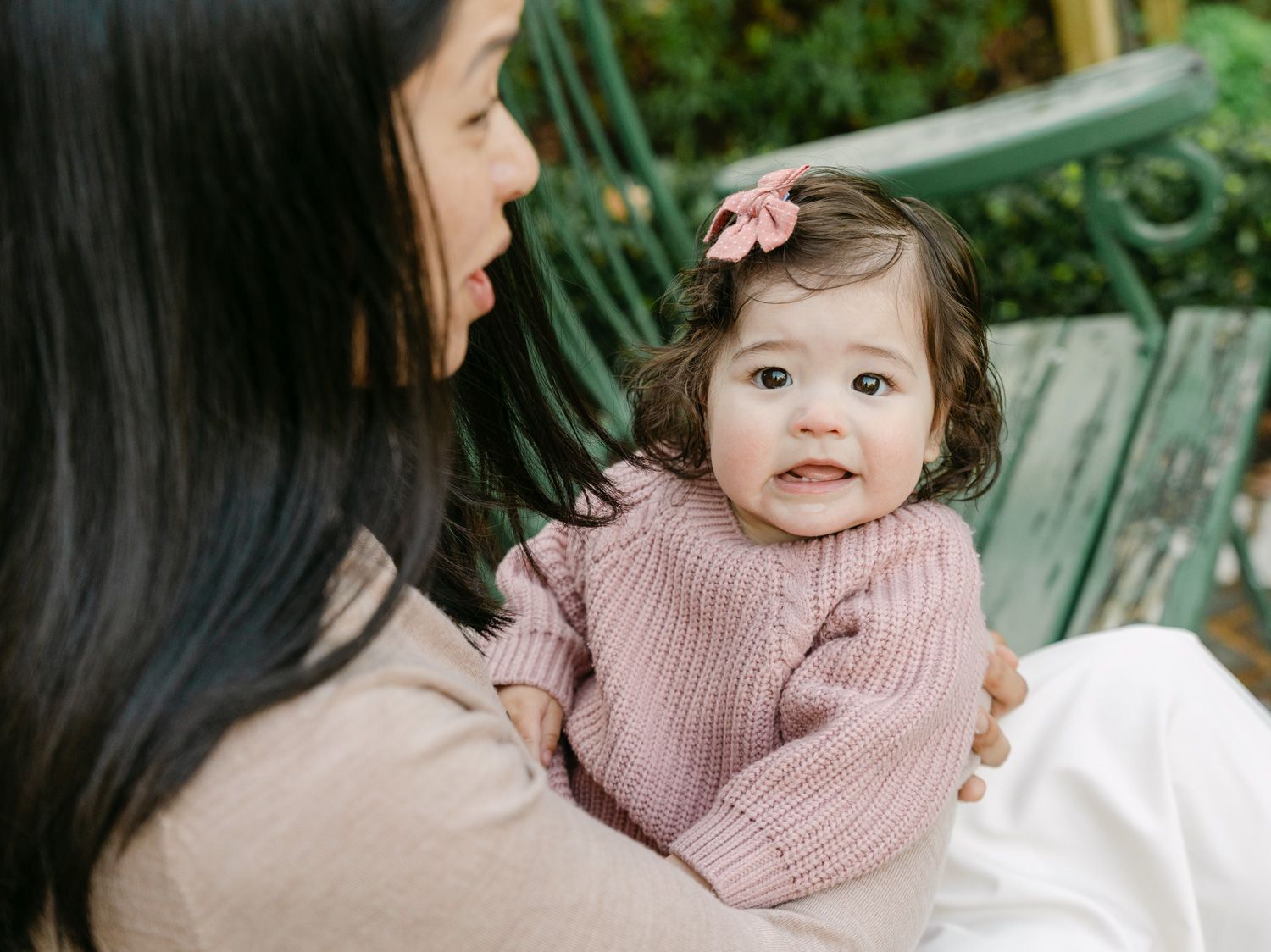 A mother holds her smiling baby girl in a pink knit sweater with a bow, sitting on a green bench outdoors.