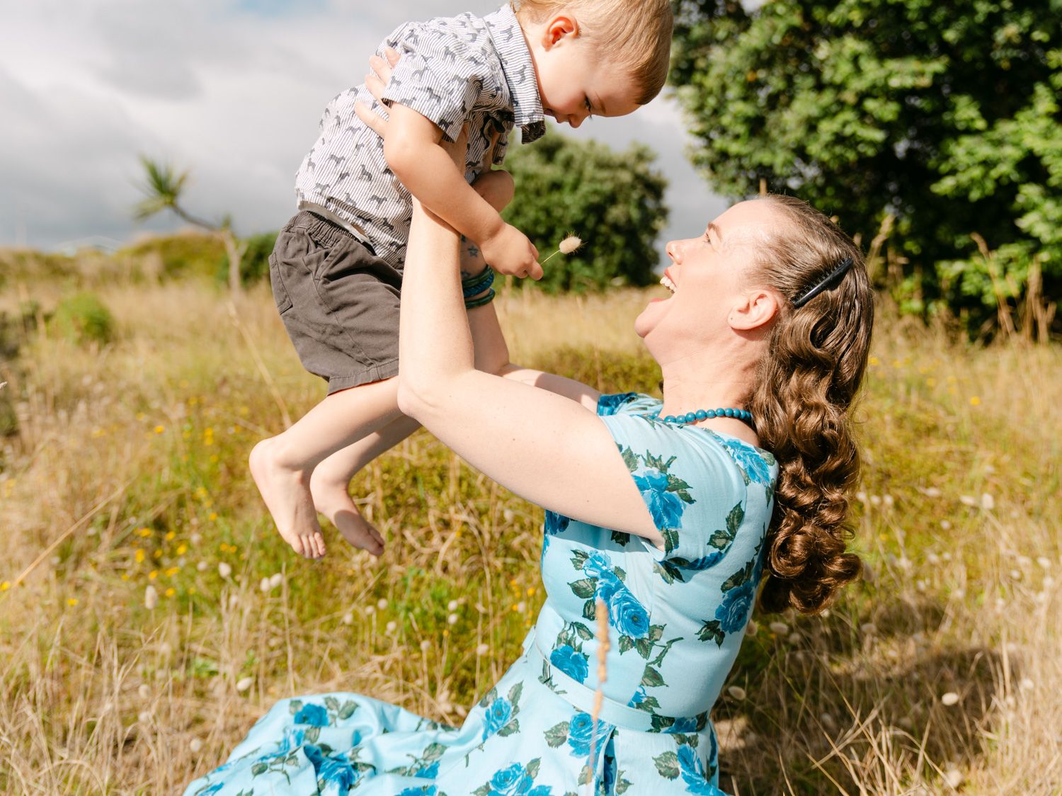 A joyful mother in a blue floral dress lifts her toddler son high in a sunny meadow.
