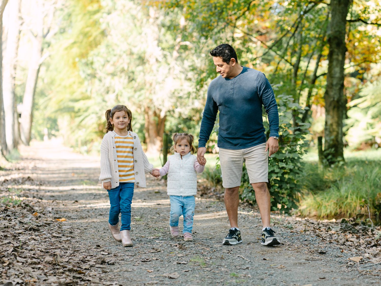 A father walks hand-in-hand with two young children on a scenic autumn park path surrounded by trees.