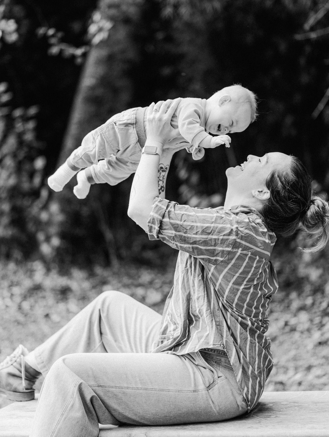 Black and white photo of a mother sitting outdoors, joyfully lifting her smiling baby up in the air.