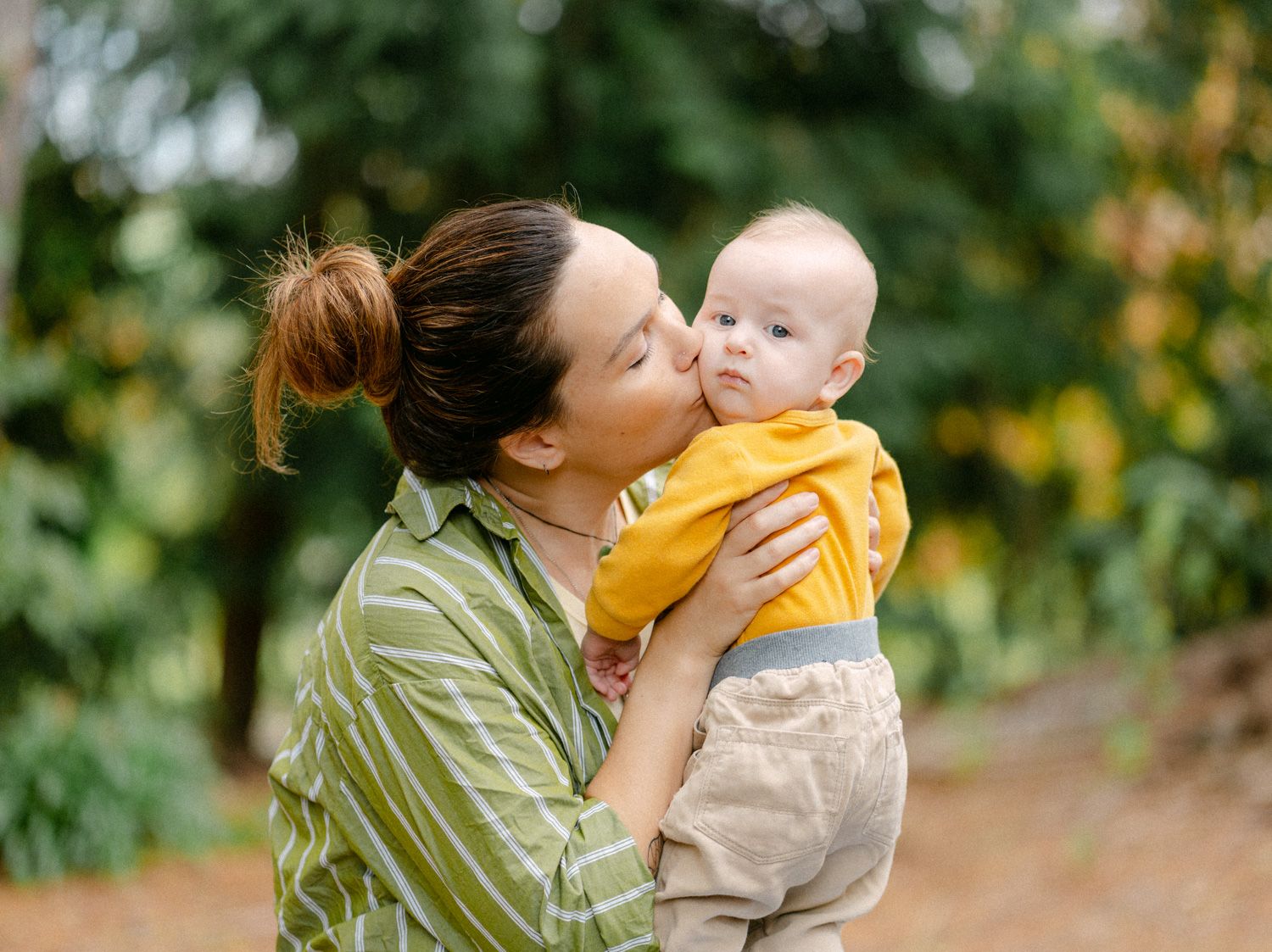 A mother tenderly kisses her smiling baby outdoors, both dressed casually amid lush green foliage.