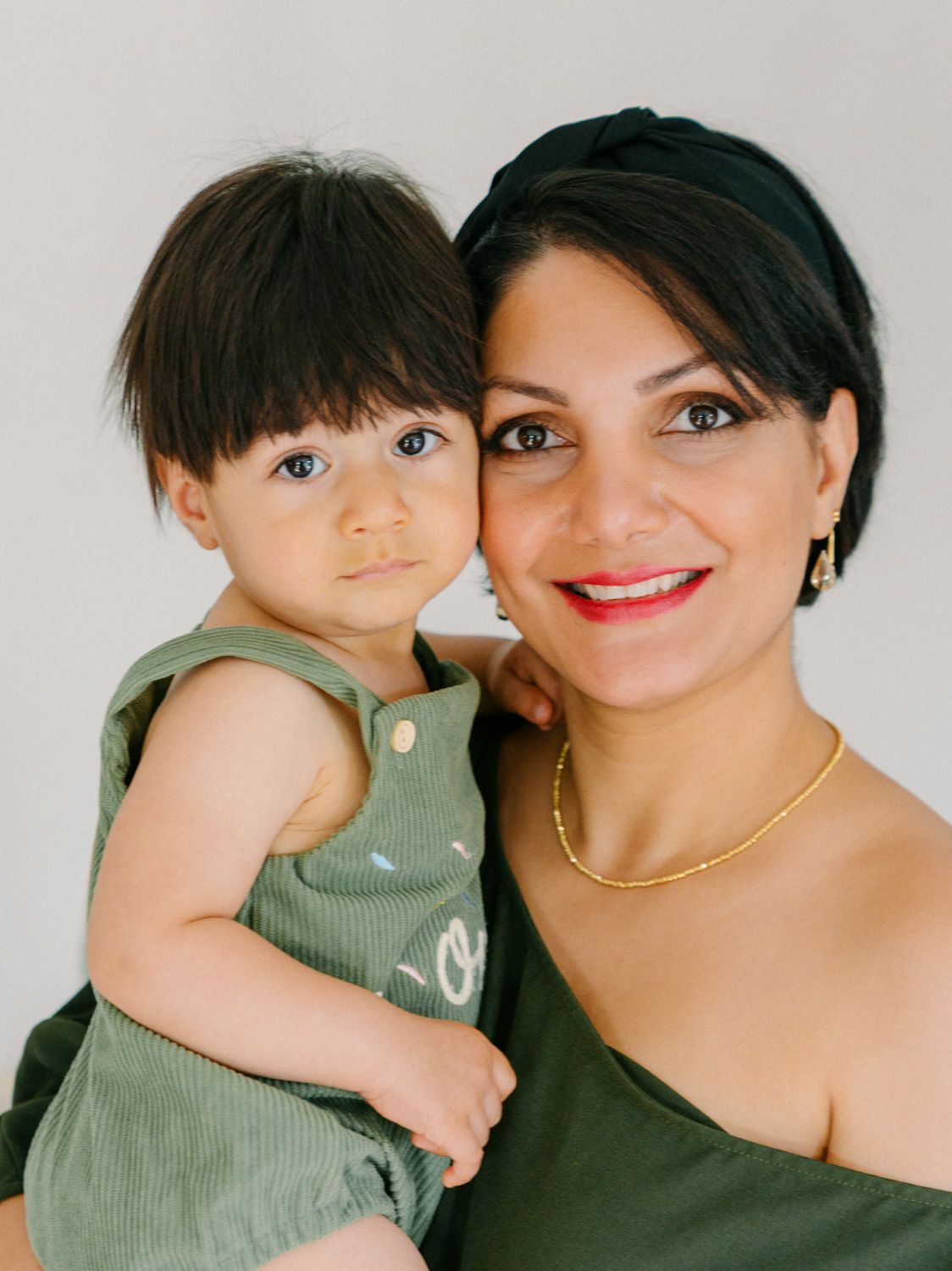 A smiling woman with red lipstick and gold jewelry holds a young toddler in a green outfit against a white background.