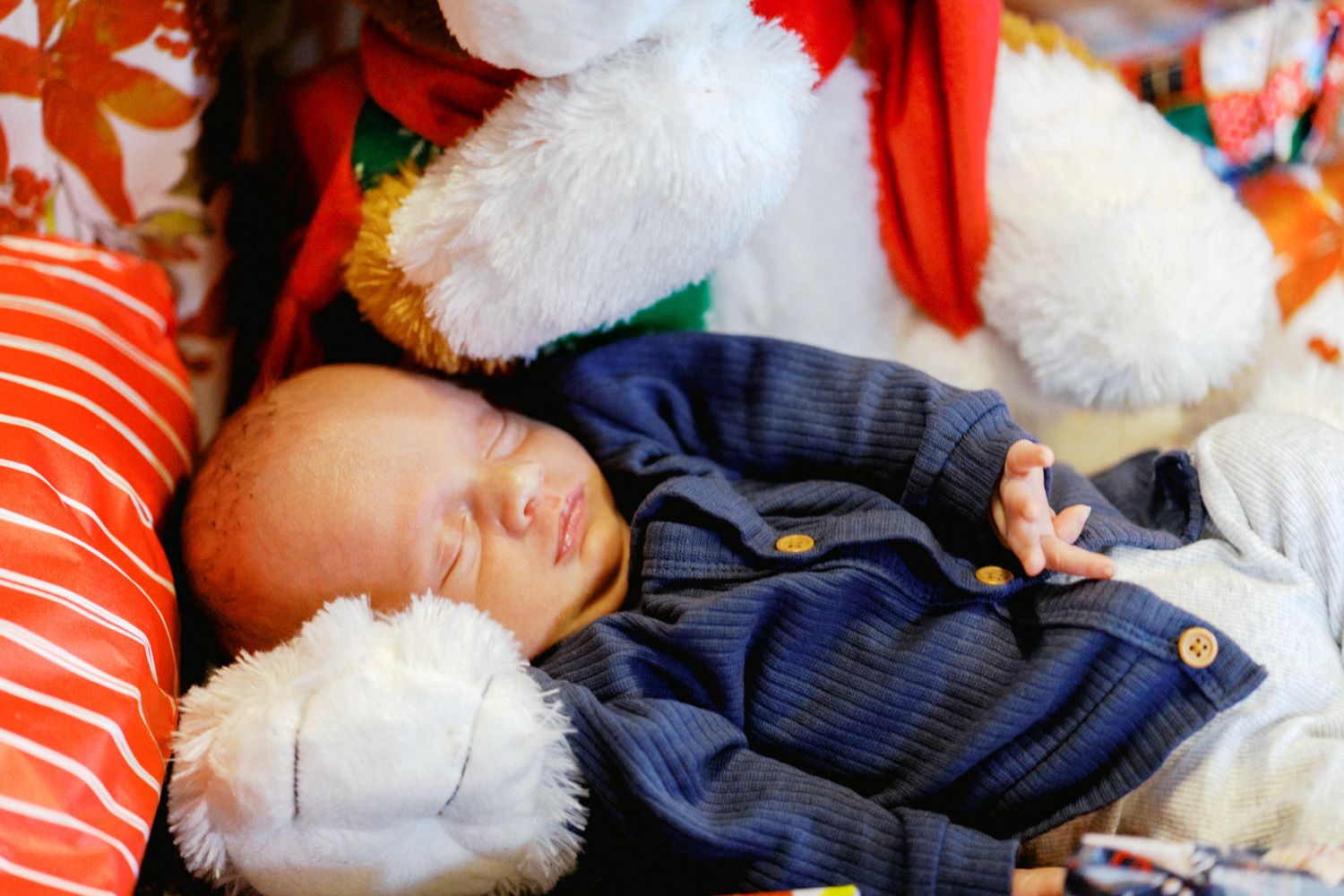 Sleeping newborn baby in blue outfit resting peacefully in Santa Claus arms during first Christmas photo.
