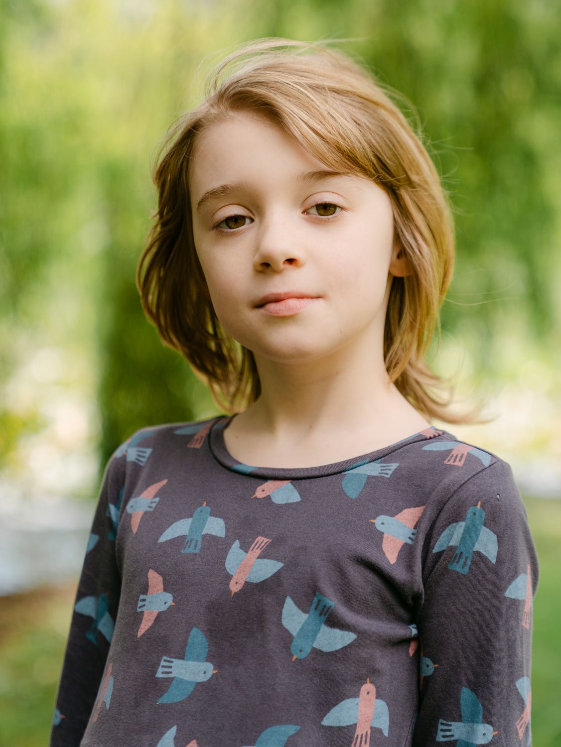 A young child with shoulder-length hair wearing a dark bird-print long-sleeve shirt, standing outdoors with green foliage behind.