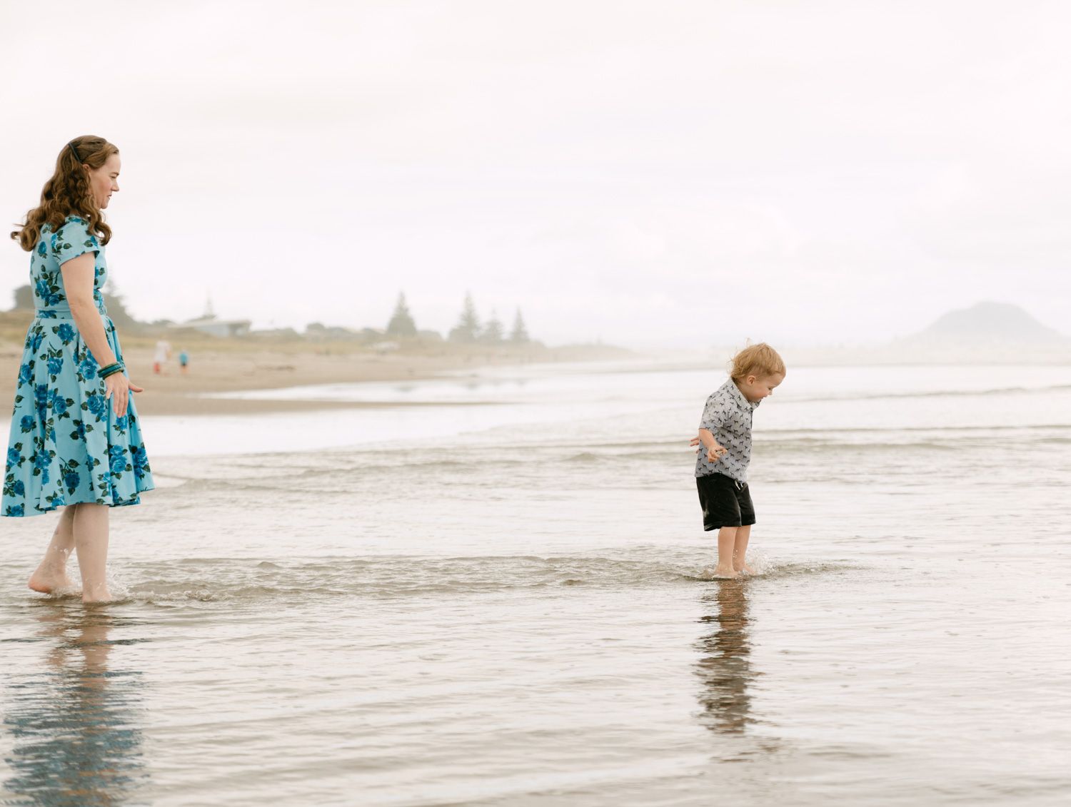 A woman in a floral dress and a child wade in shallow ocean water on a misty beach.