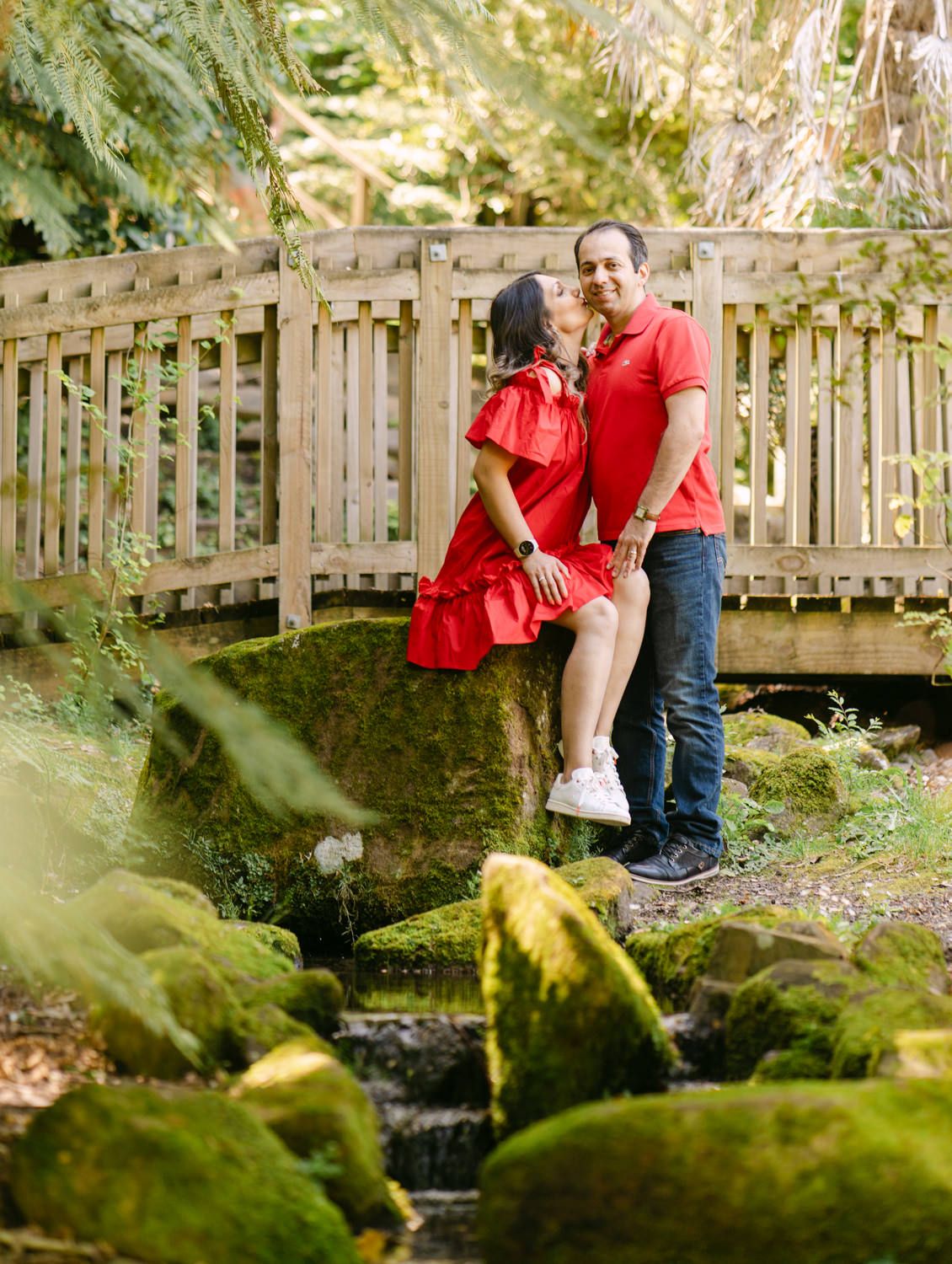 Couple in matching red outfits posing on mossy rocks near a wooden bridge in a lush green forest setting.