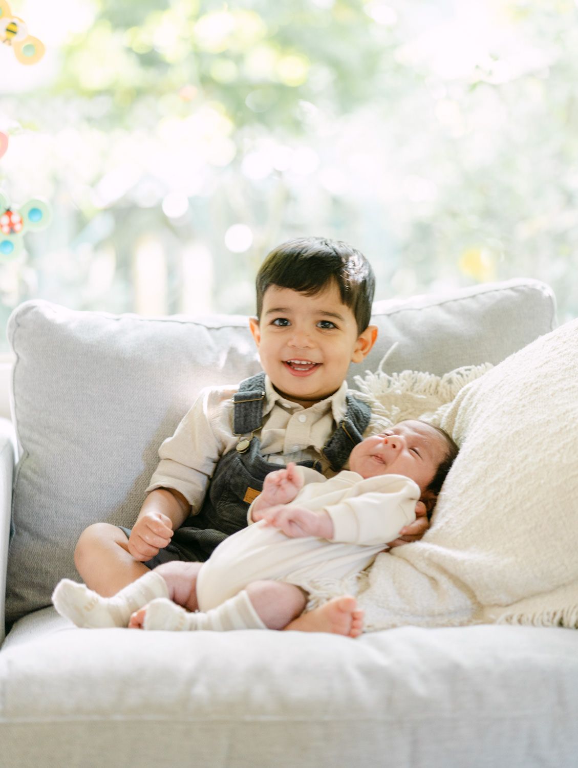 Smiling toddler boy sitting on white sofa holding newborn baby sibling in a bright, airy room.