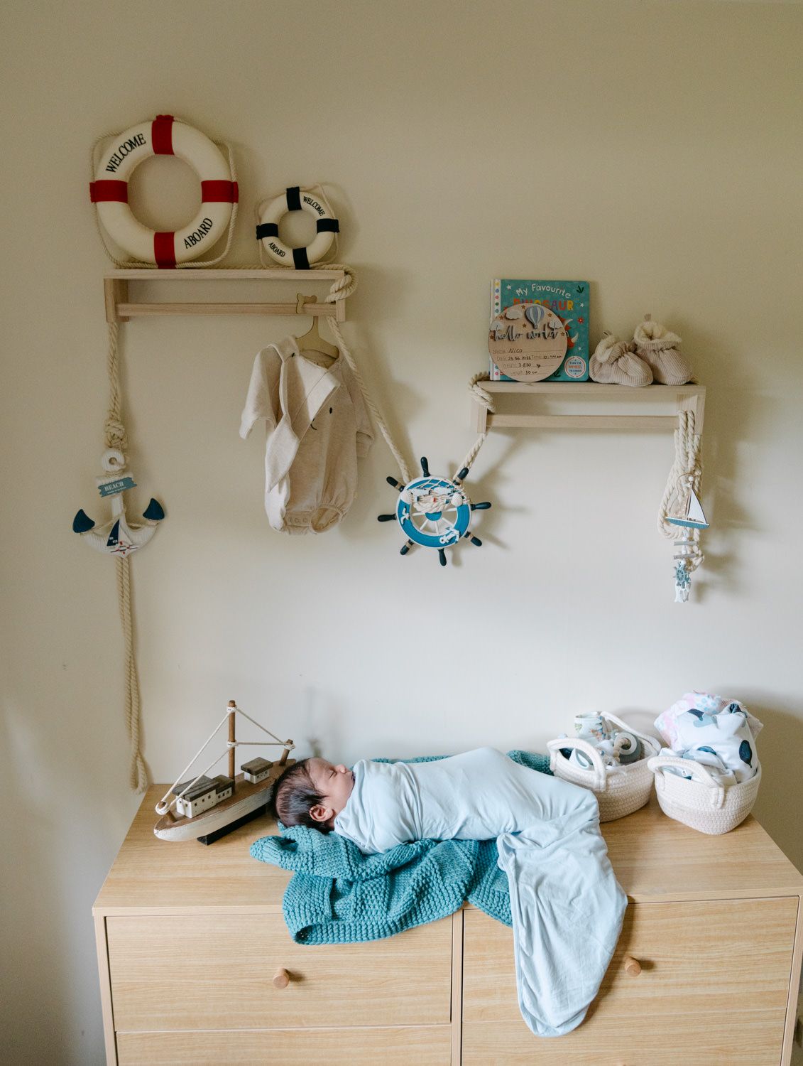 Swaddled newborn baby resting on a dresser in a nautical-themed nursery with decorative wall shelves.