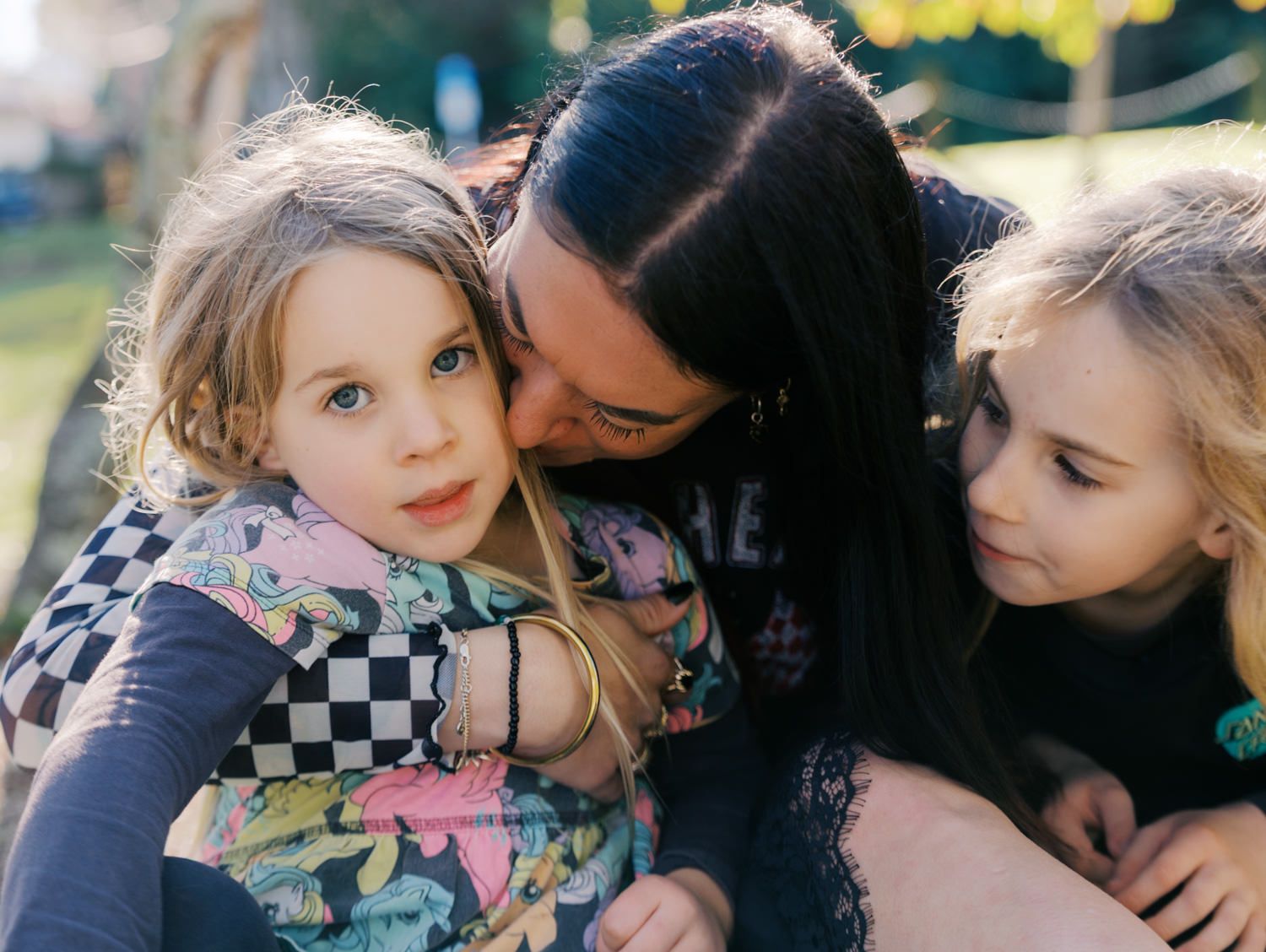 A mother tenderly embraces her two young daughters in a warm and loving outdoor close-up portrait.