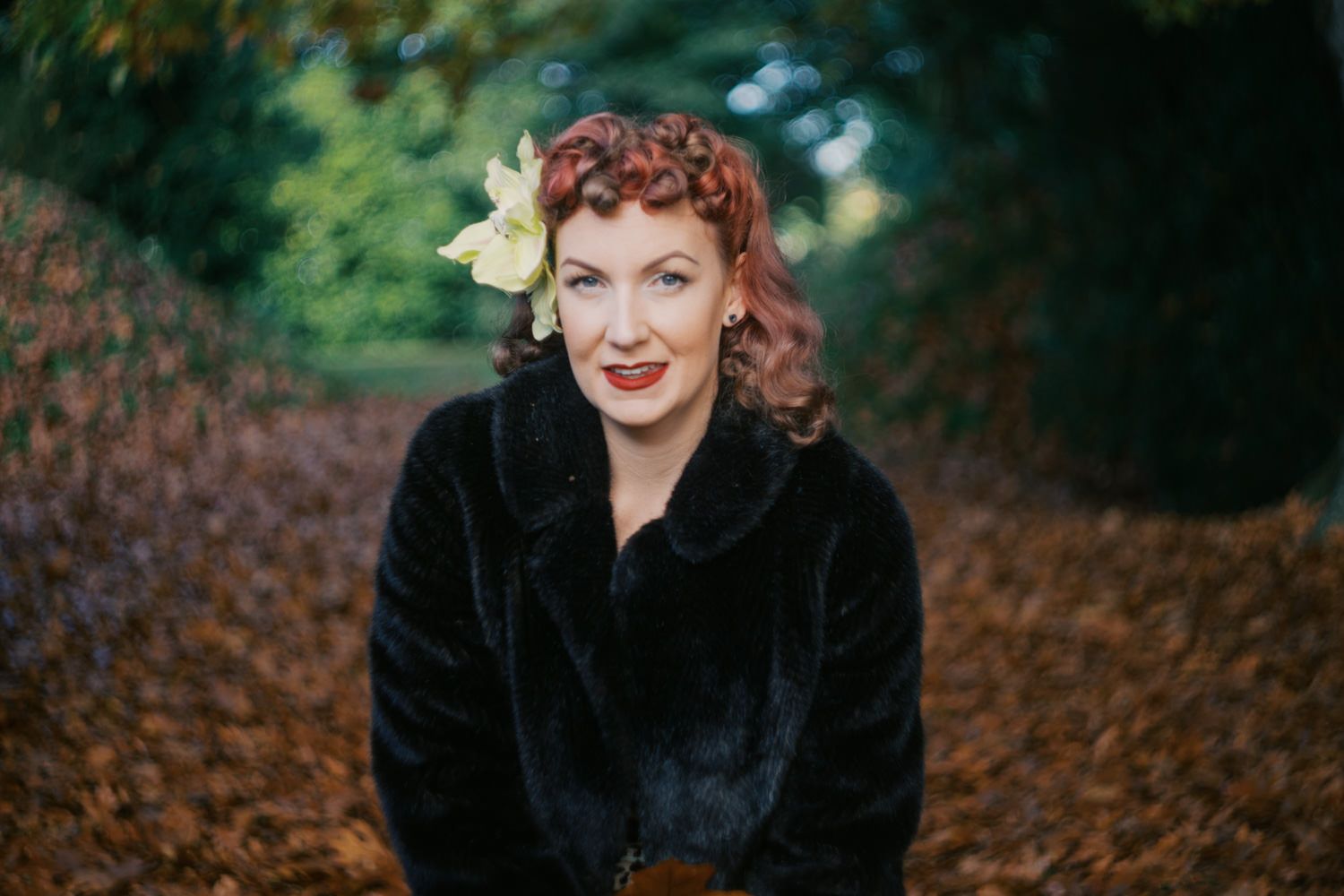 Woman in black fur coat with flower hair accessory smiling outdoors among autumn leaves.