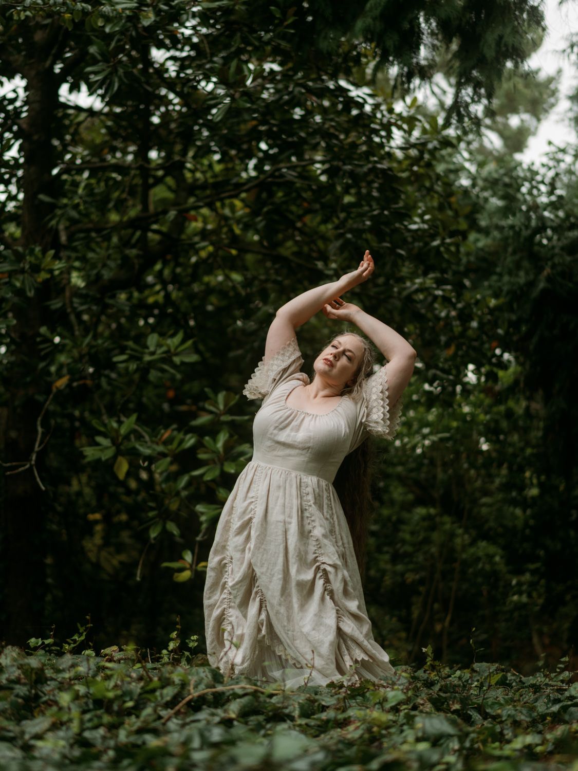 Woman in white lace dress dancing gracefully among lush green trees and ivy ground cover in a forest setting.