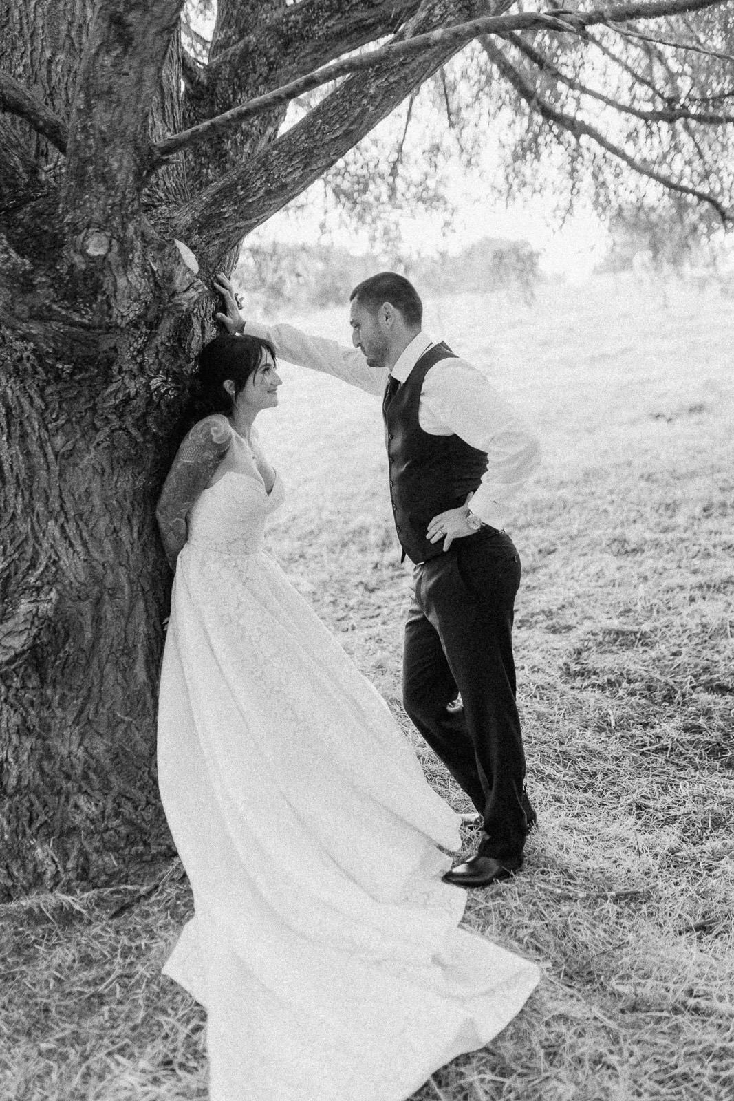 Black and white wedding photo of bride in lace gown and groom in vest leaning against a large tree outdoors.
