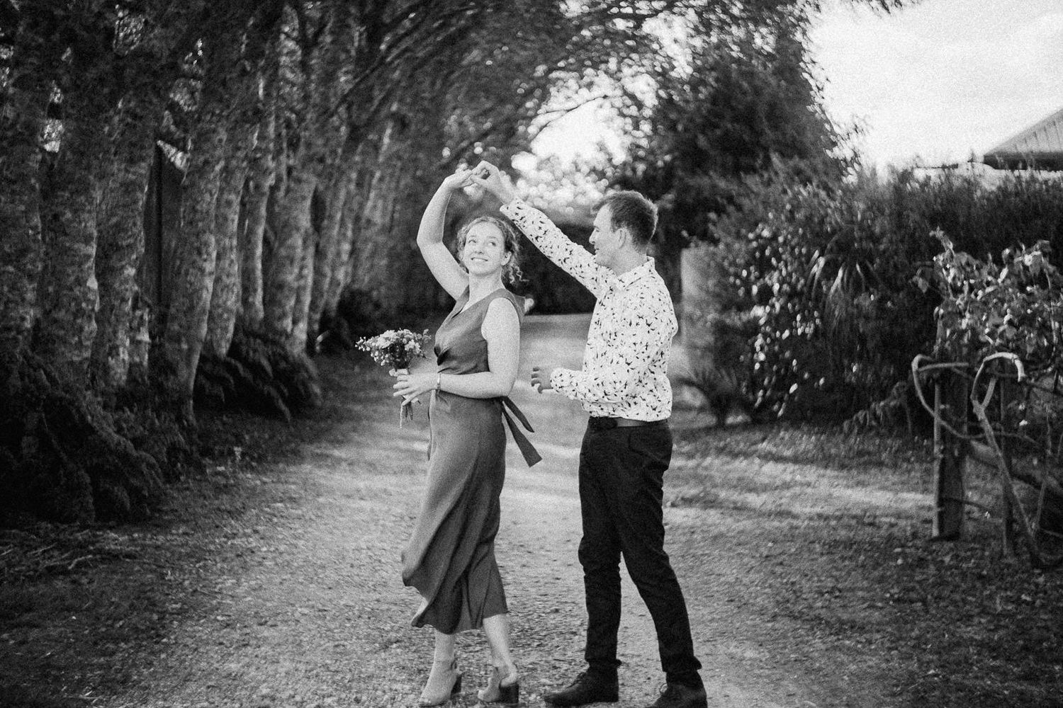 Couple dancing outdoors on a tree-lined path, man spinning woman in black and white photo.
