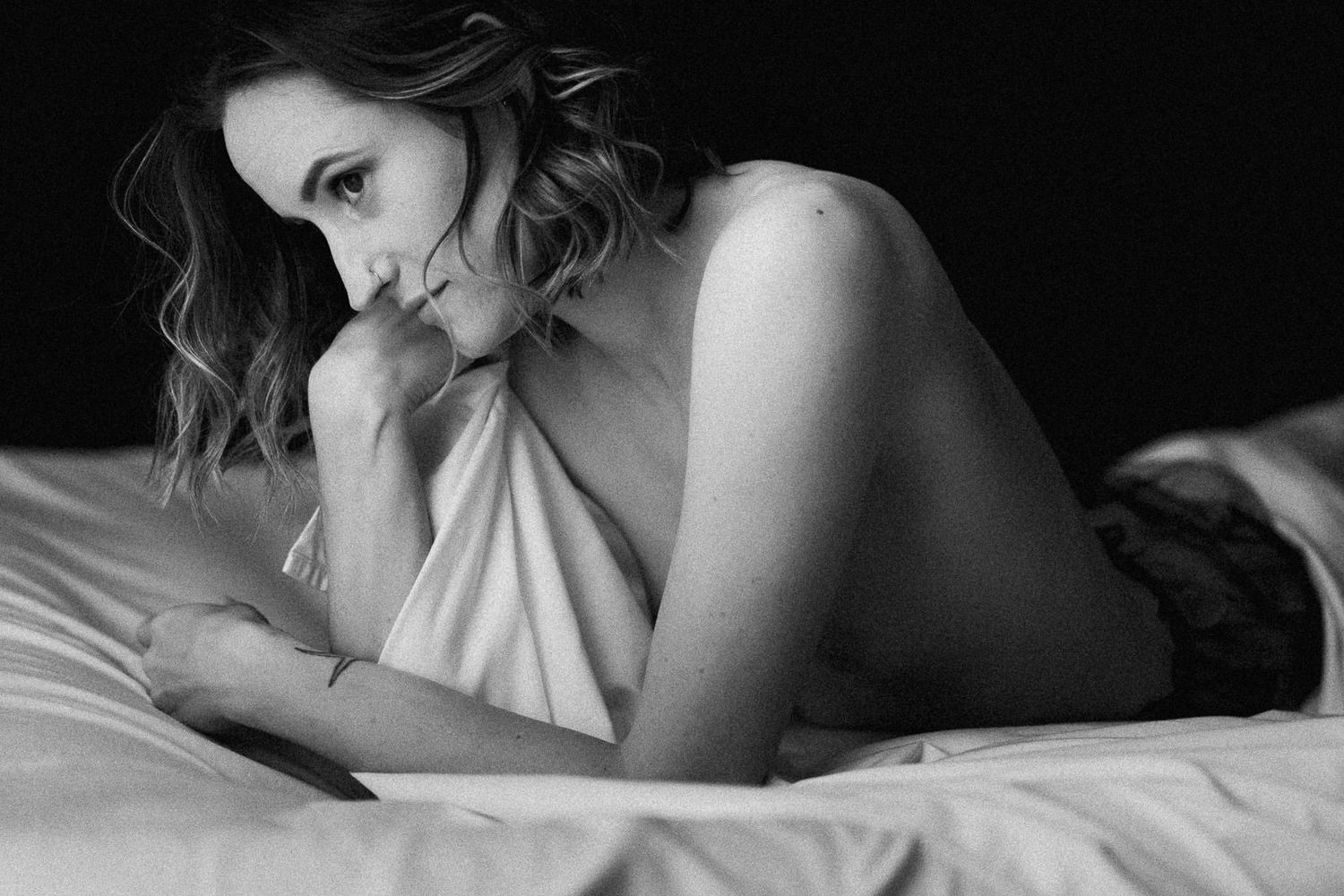 Black and white boudoir photo of a woman lying on white bed sheets in a moody, artistic portrait.