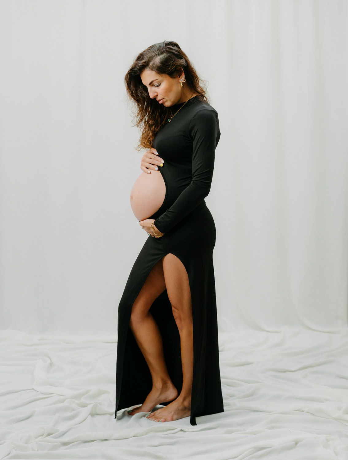 Pregnant woman in black long-sleeve slit gown cradling baby bump, studio maternity photo.