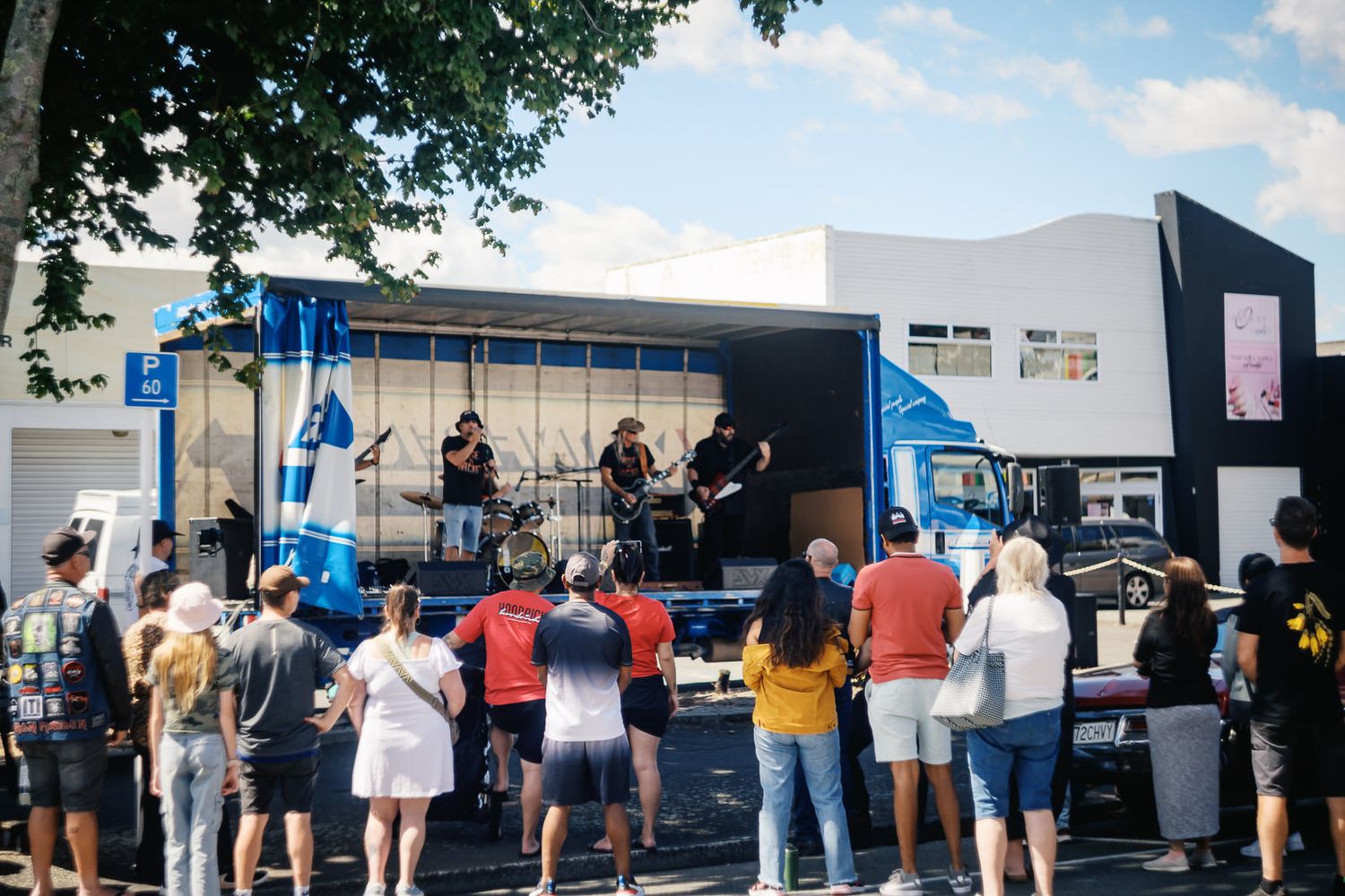 Live band performing on a mobile stage truck outdoors with a crowd of spectators watching on a sunny day.