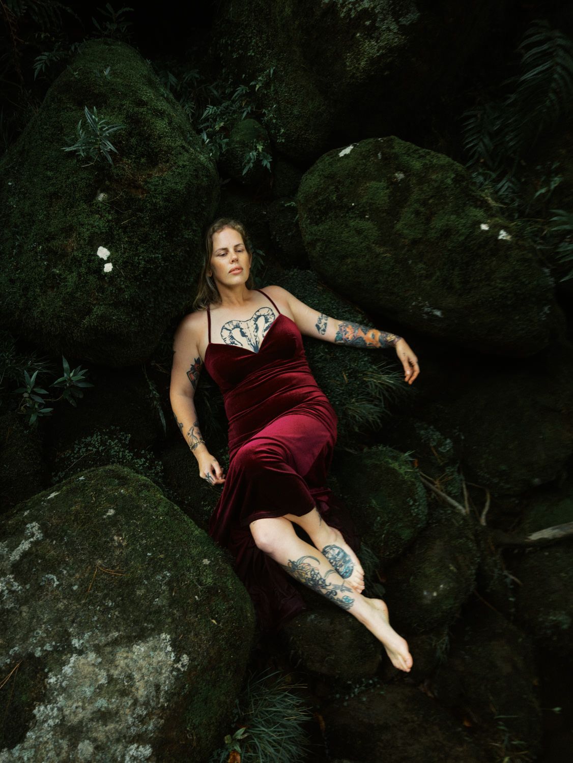 Tattooed woman in burgundy velvet slip dress reclines gracefully on moss-covered rocks surrounded by greenery.