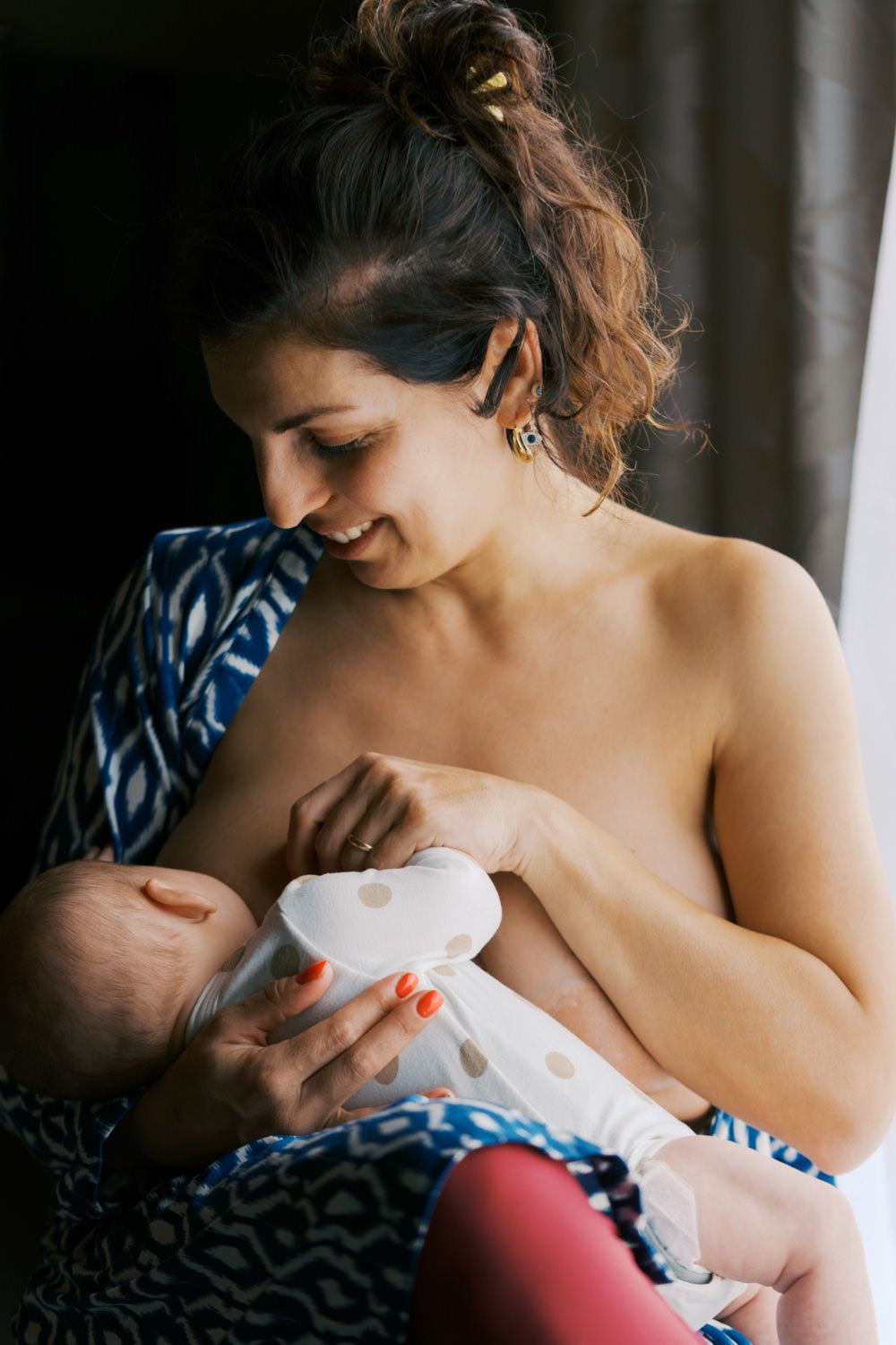 Smiling mother breastfeeding her newborn baby while sitting comfortably, wearing a blue patterned wrap.