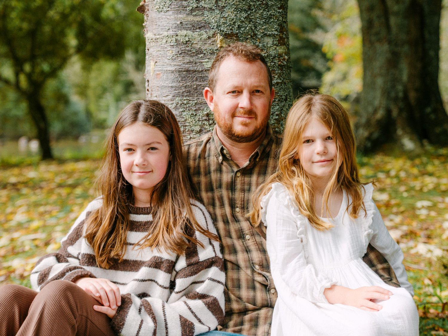 A father poses with two young daughters under a tree in an autumn park setting.