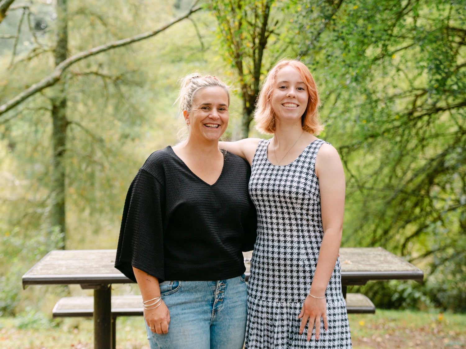 Two women smiling together outdoors in a lush green forest setting, one in black top with jeans, other in checkered dress.