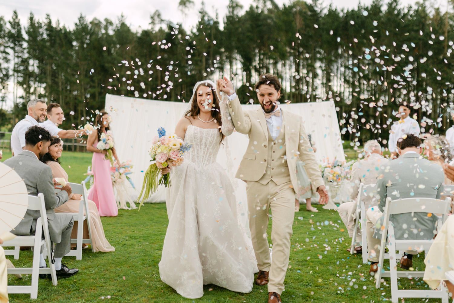 Bride and groom walk joyfully through confetti shower at outdoor wedding ceremony on lush green lawn.