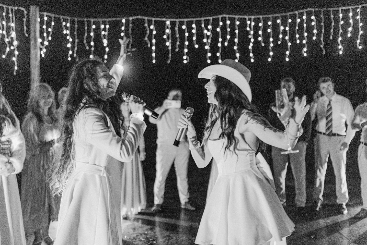 Two brides in white dancing together under fairy lights at wedding reception in black and white photo.