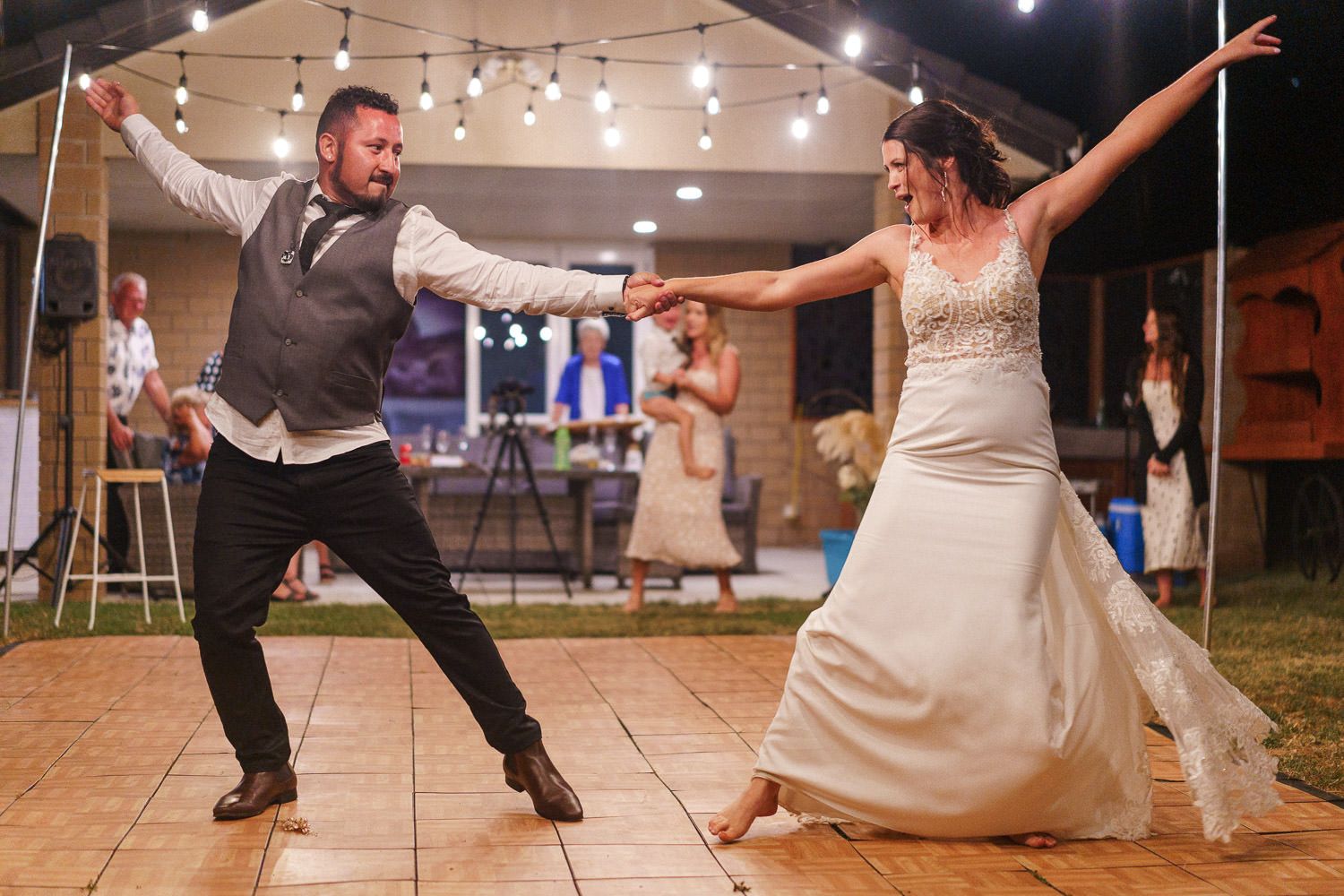 Bride and groom share energetic first dance outdoors at evening wedding reception with string lights.