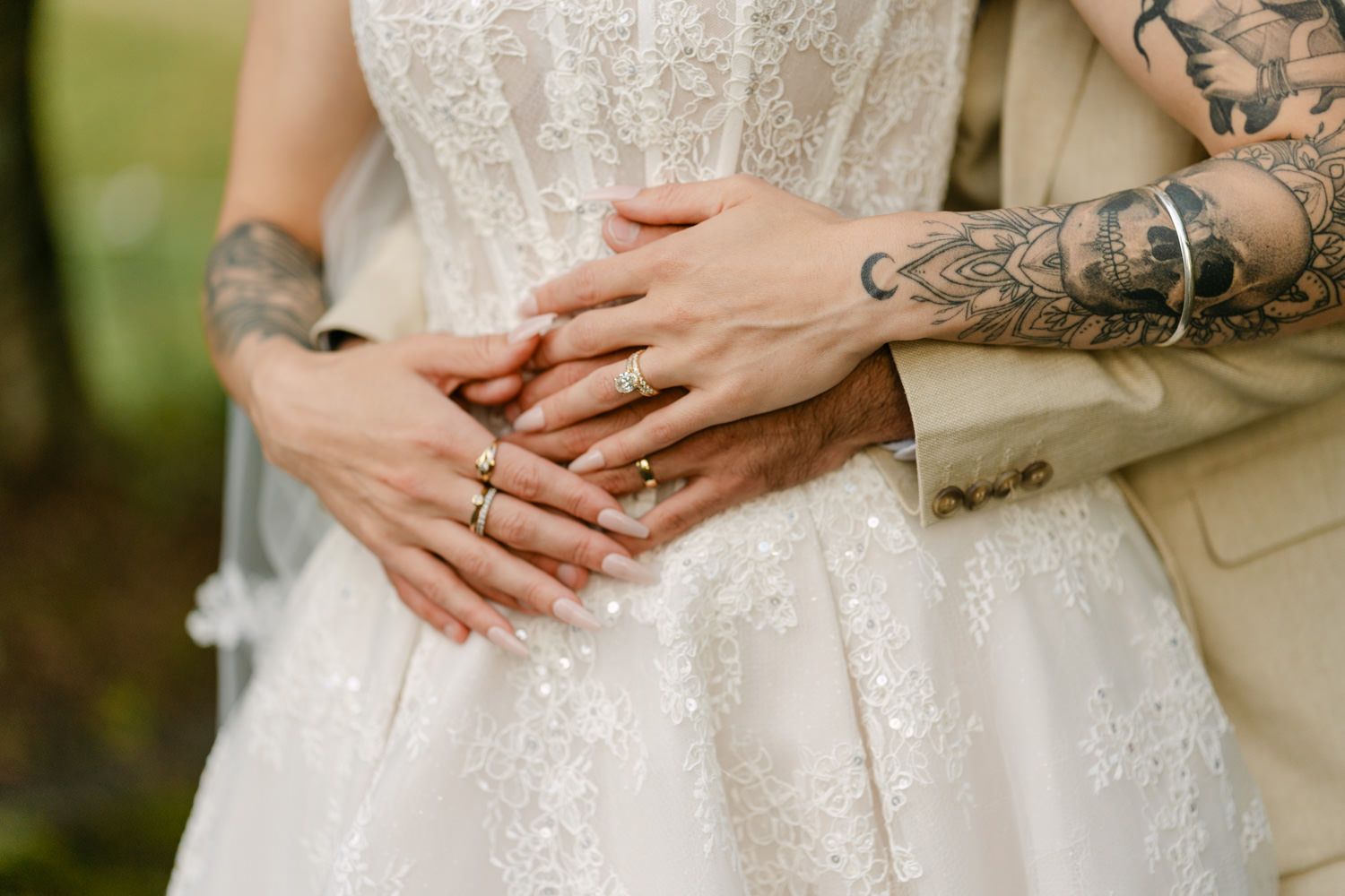 Tattooed couple in wedding attire with gold rings, bride in lace dress, groom in beige suit, hands resting on her waist.