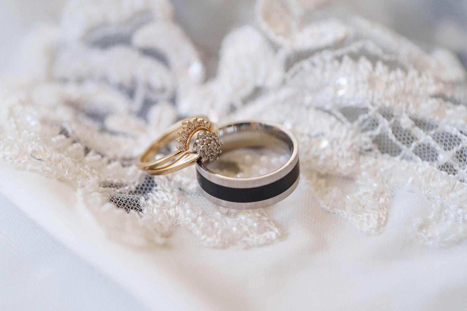 Two wedding rings, a gold diamond ring and a black and gold band, resting on white lace fabric.