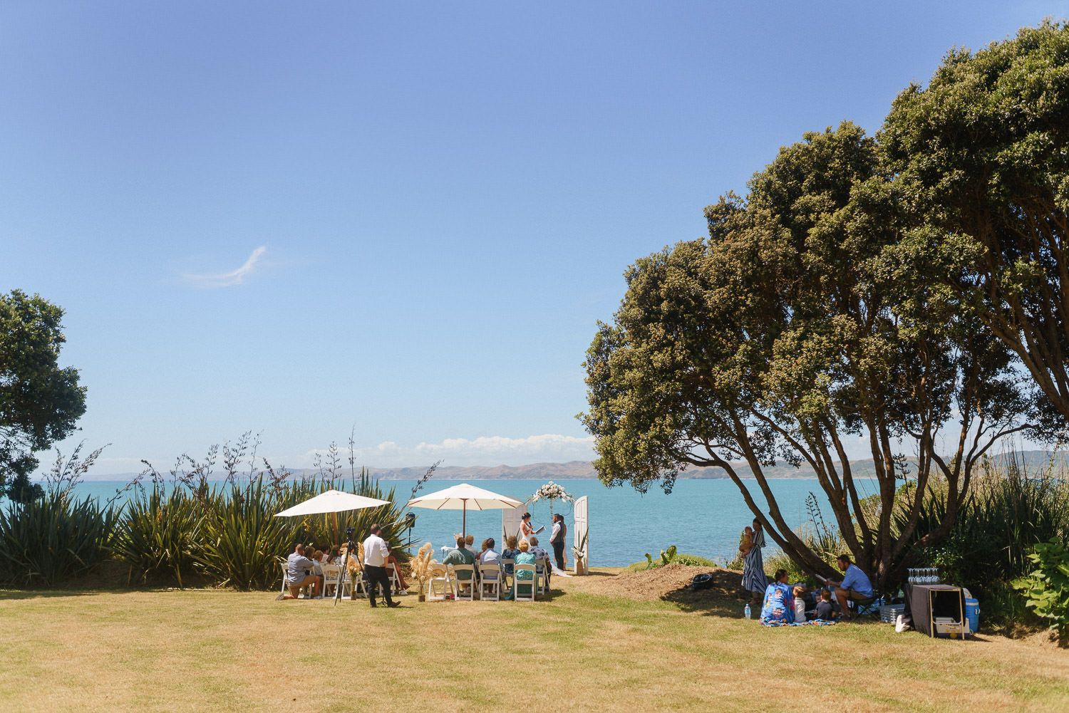 Outdoor coastal gathering on green lawn with umbrella, trees, and stunning blue ocean views on a sunny day.